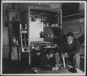 Black and white photograph of two men looking at blueprints. They are seated in front of a large metal apparatus.