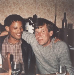 Colour photograph of John Foster and Juan Cespedes sitting side by side with their arms around each other.