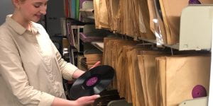Woman holding a '78 inch vinyl record, in front of shelf full of vinyl records in paper sleeves
