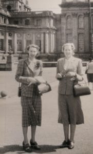 Sepia photo of two women in period dress holding handbags in front of building.