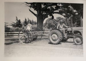 Man and child on tractor in front of large tree behind fence.